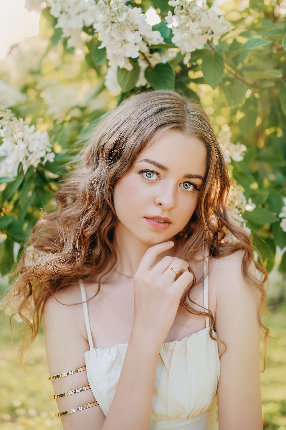 Young woman with wavy hair in a yellow dress poses among white flowering lilac bushes in soft sunlight.