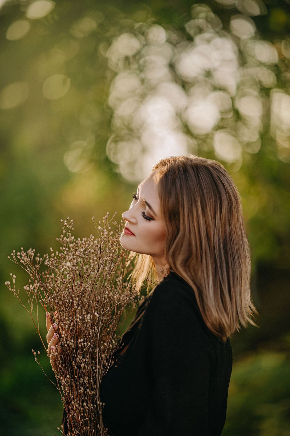 A blonde woman in a black jacket holds dried wildflowers, posed in a sunlit garden with soft bokeh background.