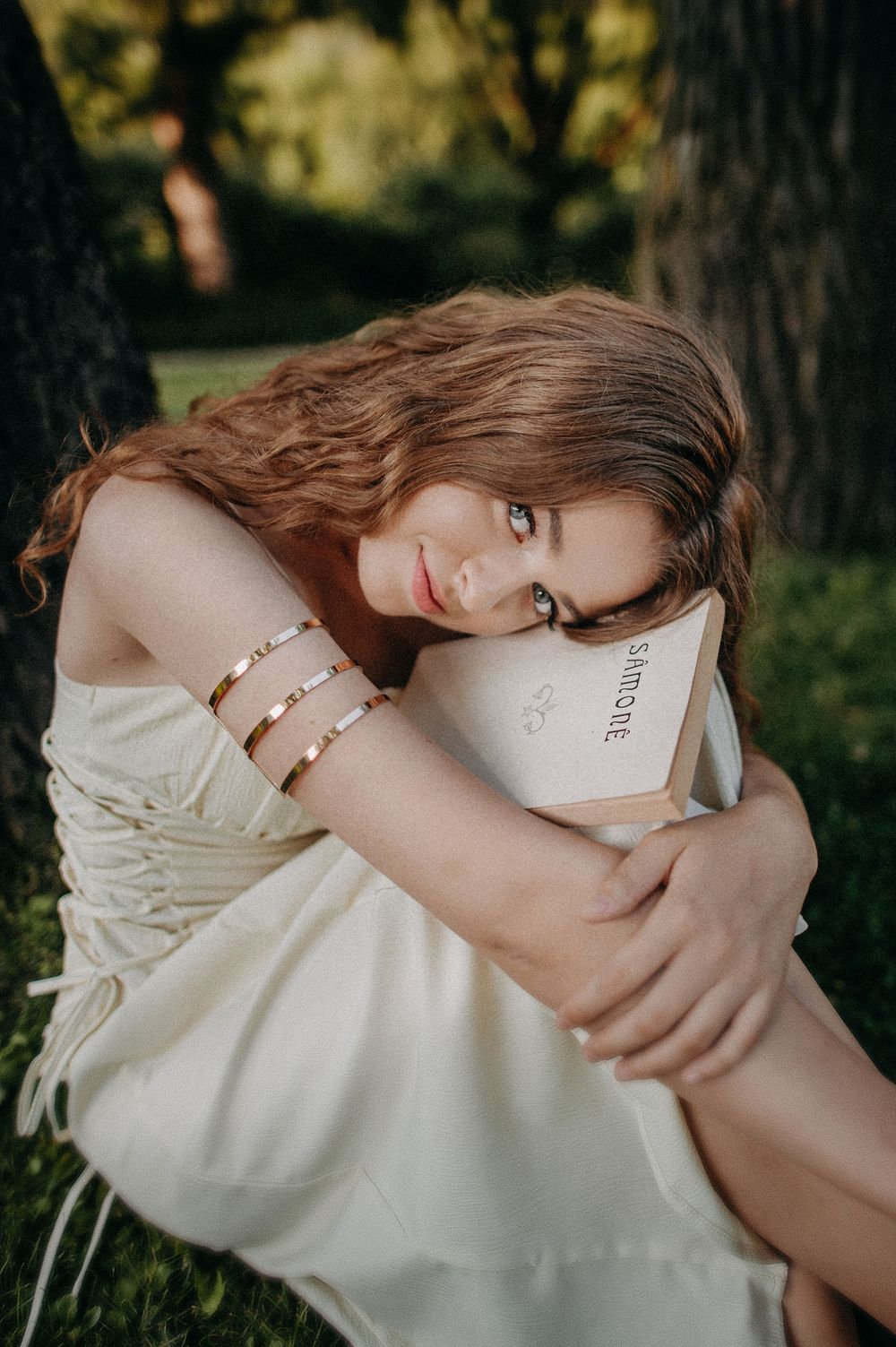 Young woman with auburn hair in white dress hugging a book while sitting against a tree outdoors.