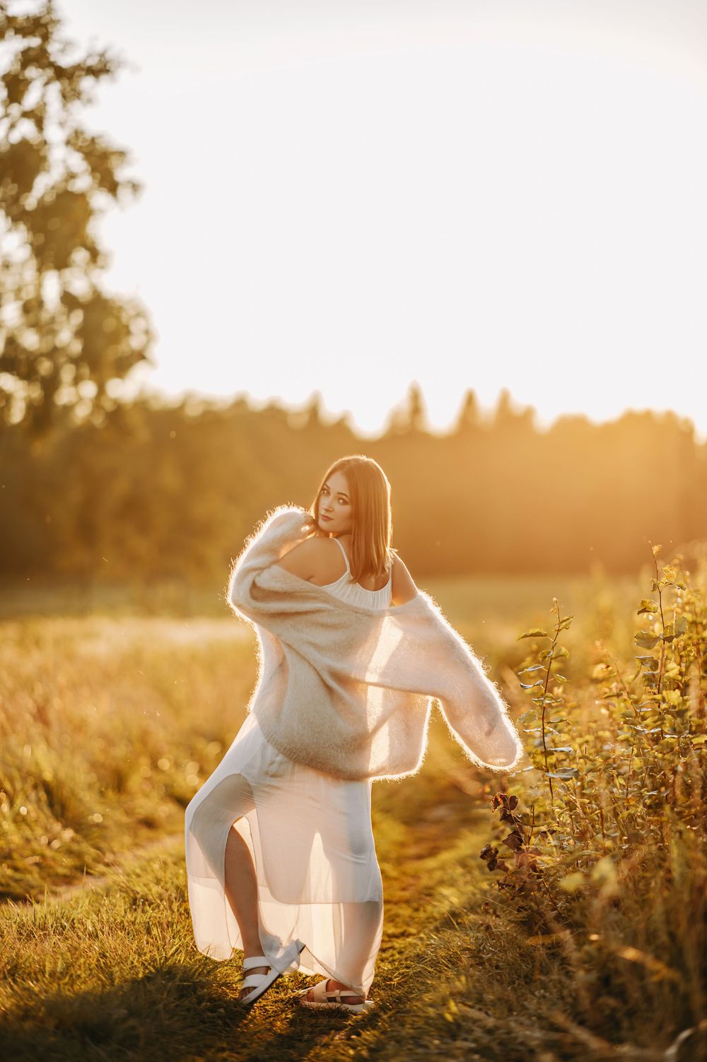 Woman in white dress and cardigan posing in golden sunset light in a field with trees in background.