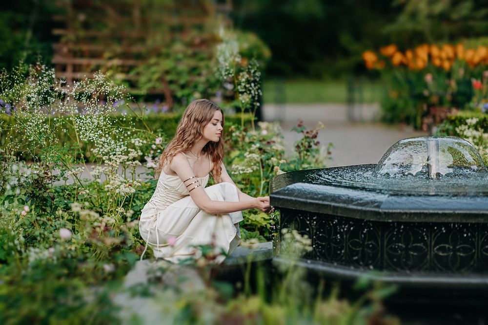 Young woman in white dress kneeling by ornate garden fountain surrounded by colorful blooming flowers.