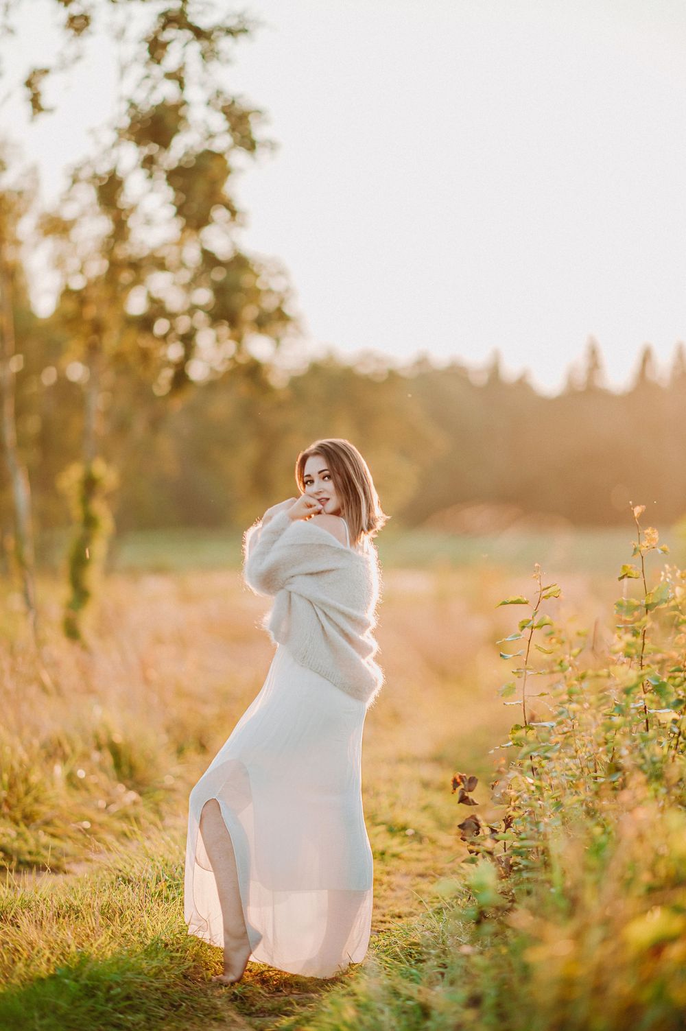 Woman in white dress and oversized sweater poses in golden hour sunlight amid lush greenery.