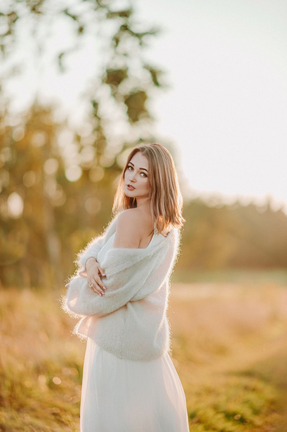 Young woman in white dress and fluffy wrap poses outdoors in golden hour sunlight with bokeh forest background.