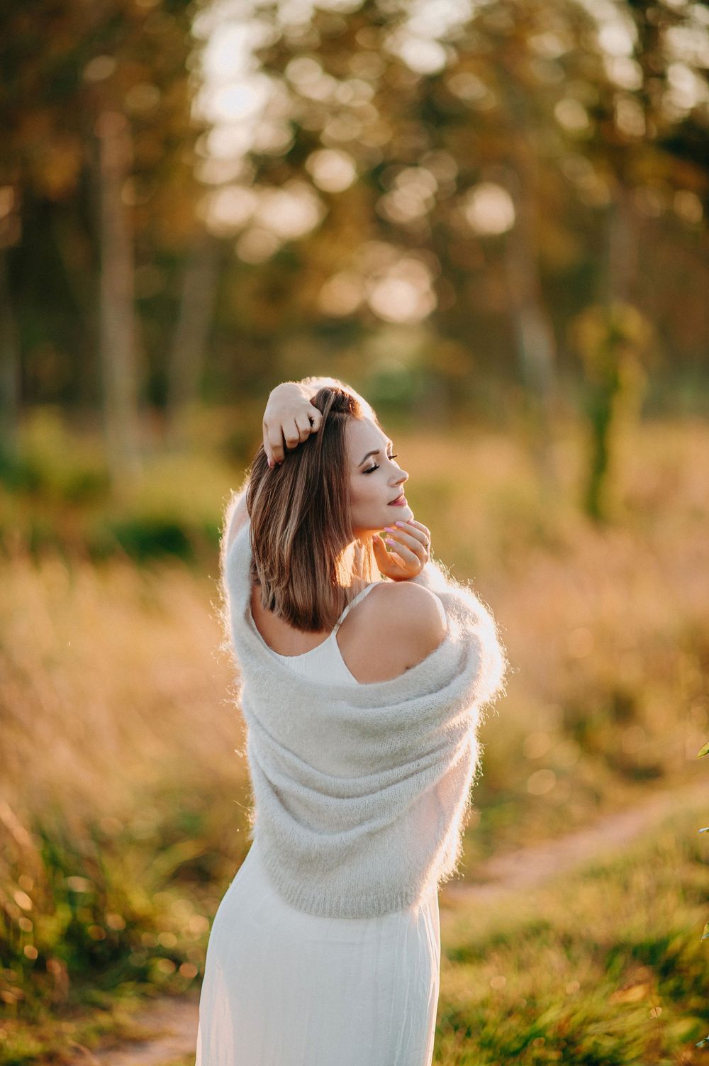 A woman in a white dress and fluffy sweater poses gracefully outdoors in golden autumn light.