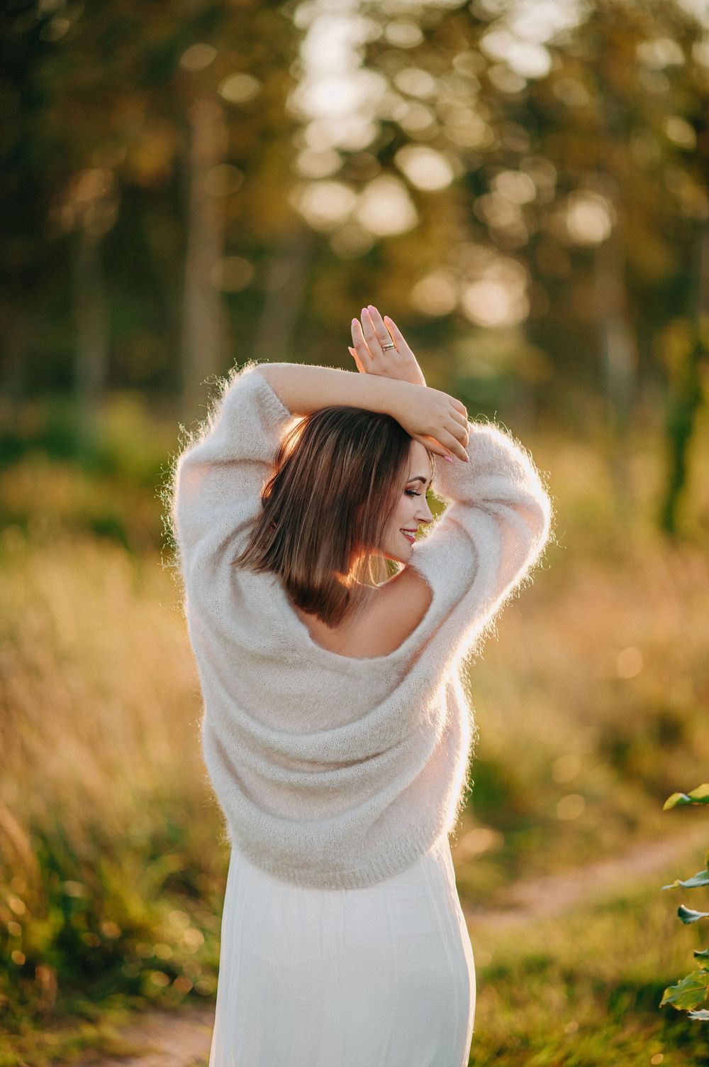 Woman in white fuzzy sweater poses with arms raised outdoors in golden hour sunlight near a forest path.