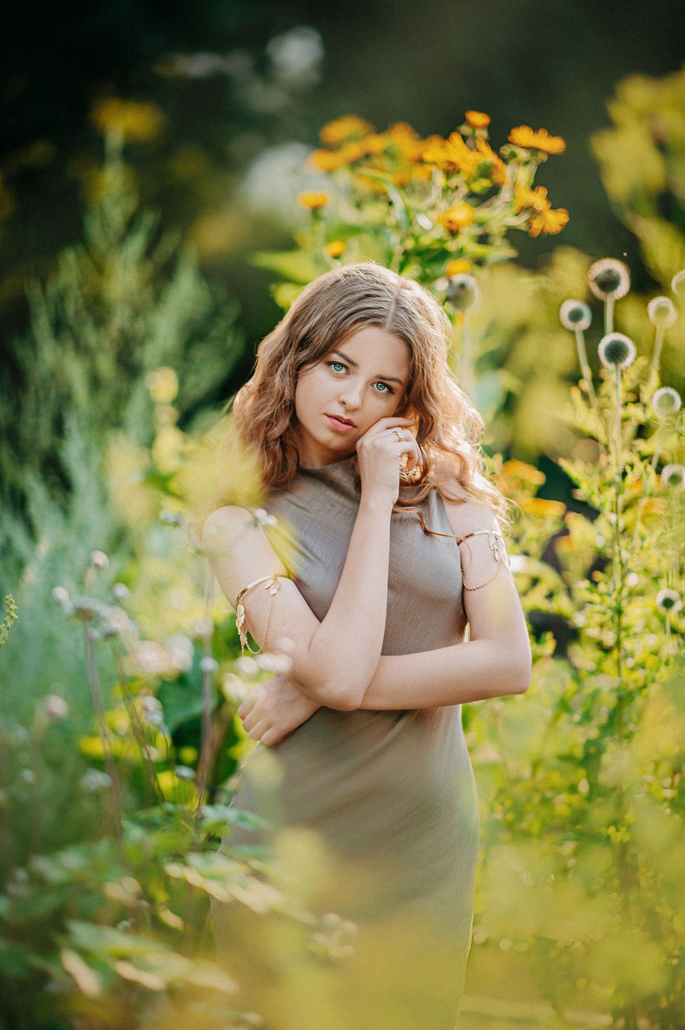 Young woman with curly blonde hair in beige dress surrounded by yellow wildflowers in a sunlit garden.