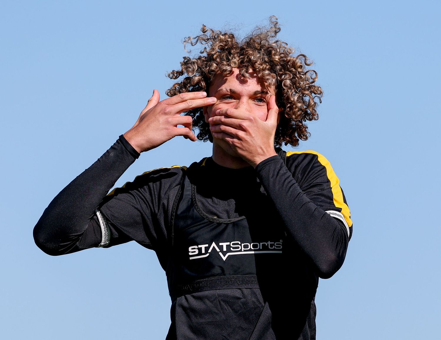 Curly-haired athlete in STATSports black training vest covering mouth with hands against blue sky.