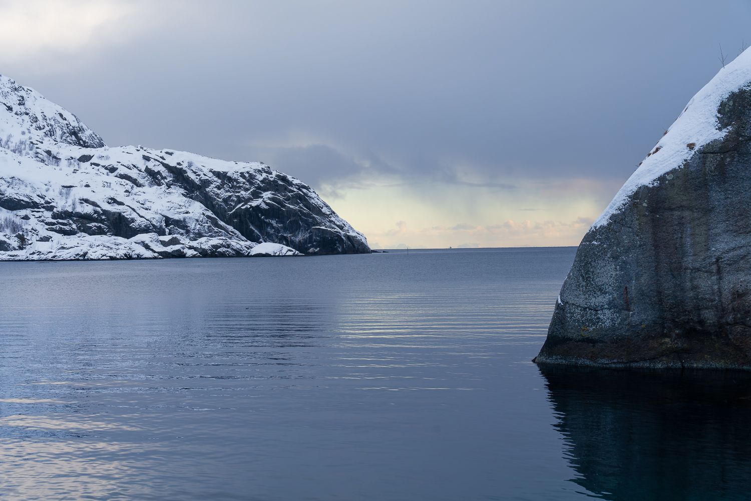 Snow-covered rocky cliffs framing a calm Arctic fjord under a cloudy winter sky.