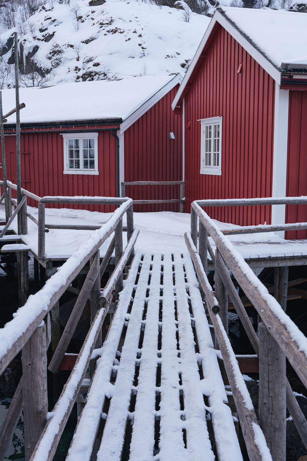 Snow-covered wooden bridge leading to a red Norwegian rorbu cabin in winter.