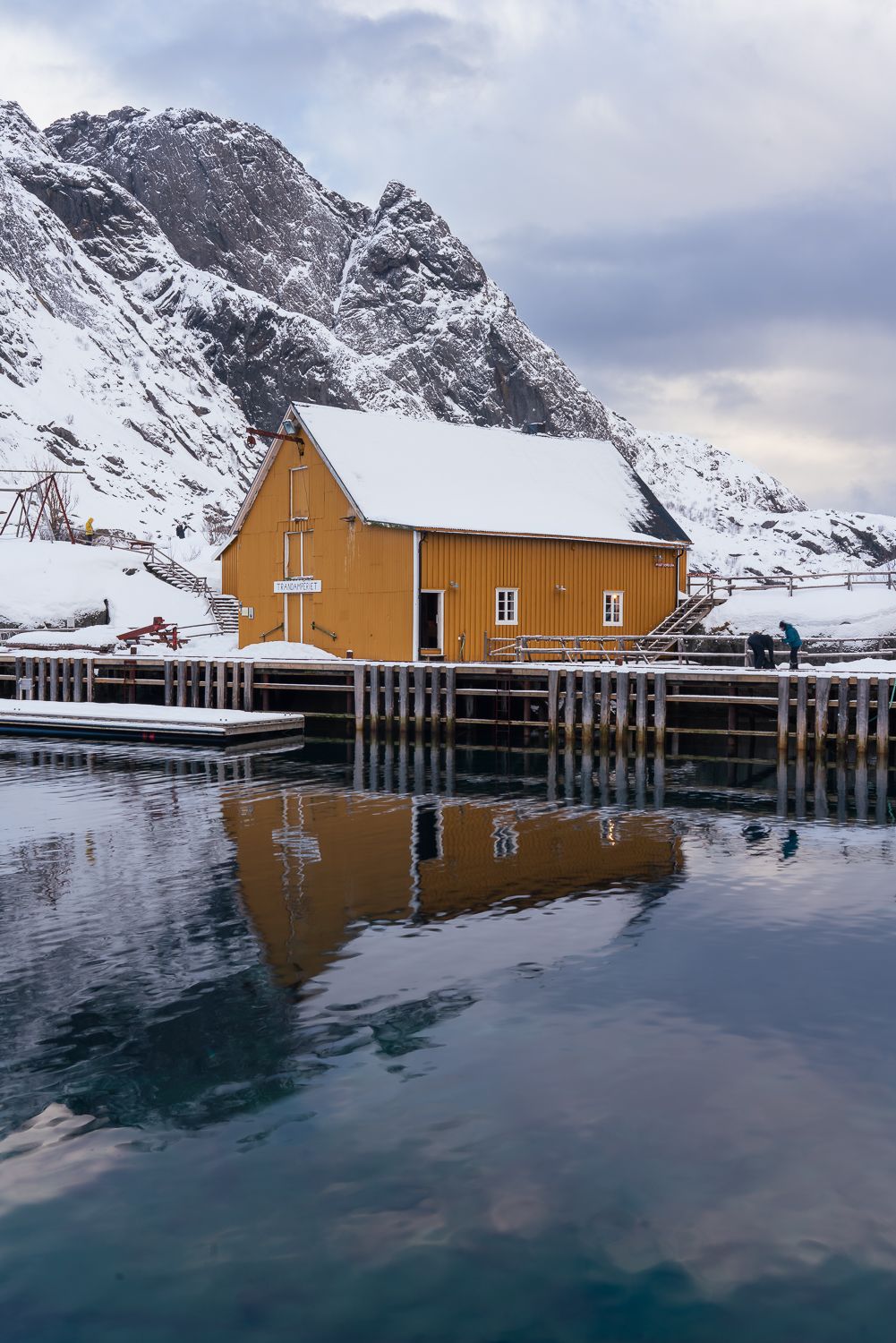 Yellow Norwegian boathouse reflected in calm fjord waters with snowy mountains behind.