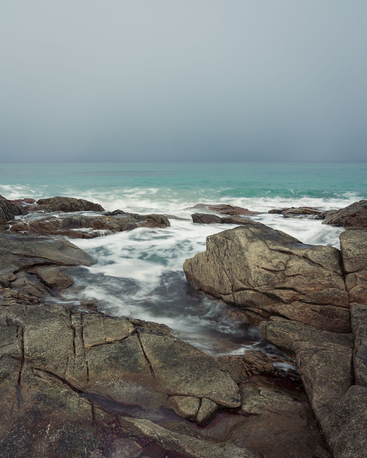 Long exposure of misty turquoise ocean waves washing over rugged rocky coastal formations under overcast skies.