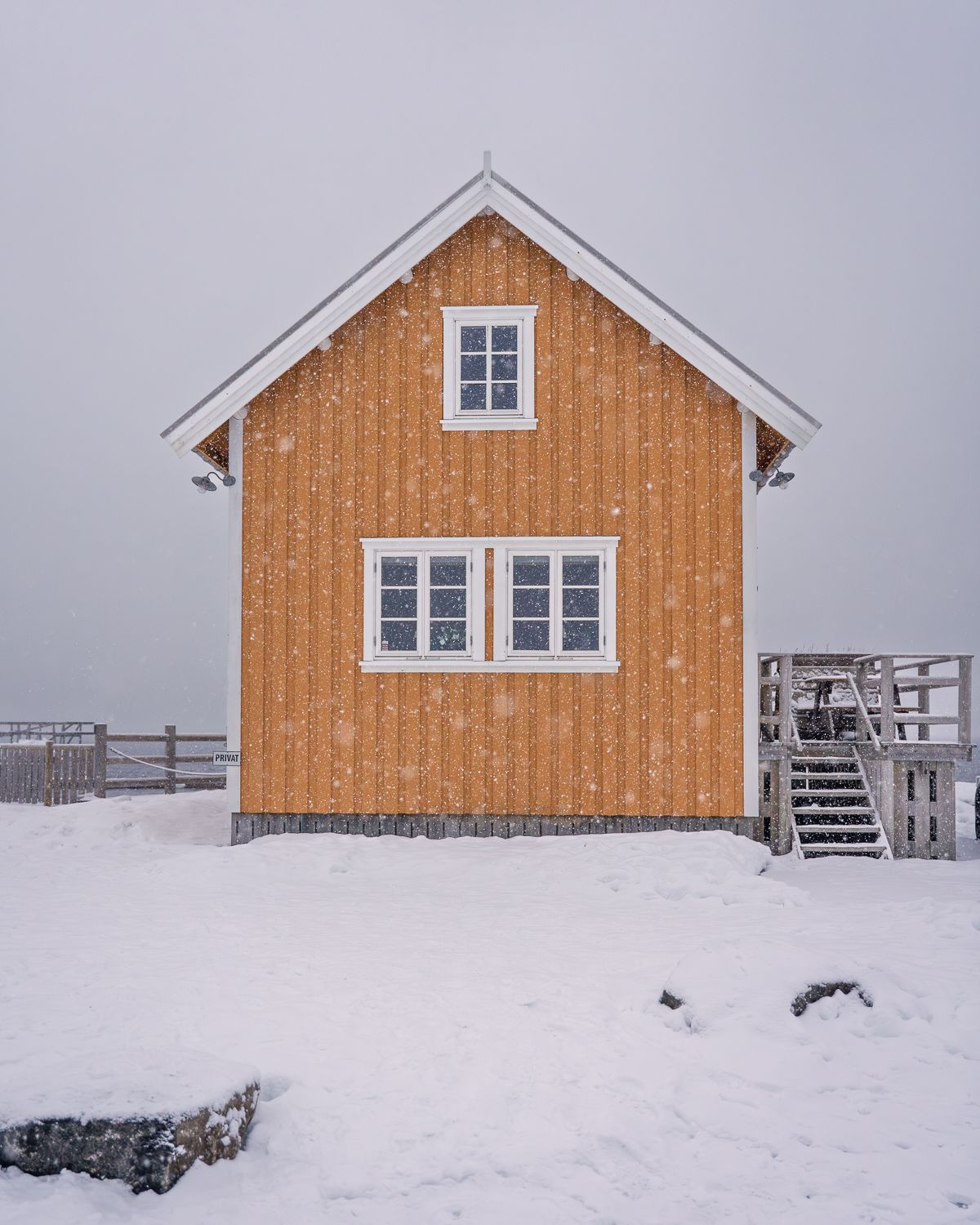 Yellow wooden Norwegian cabin dusted with falling snow stands isolated in a stark white winter landscape.