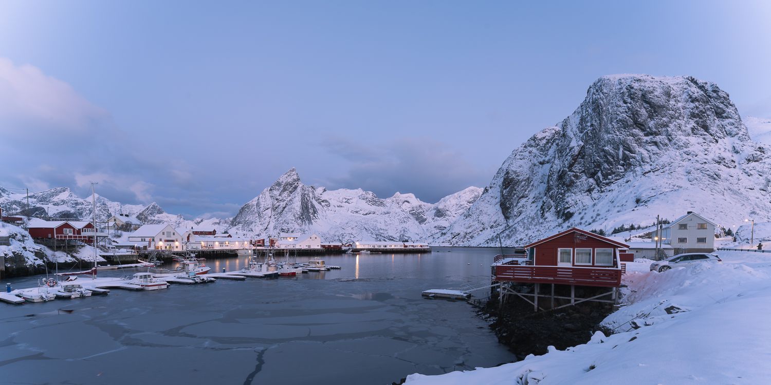 Panoramic winter view of frozen harbor with red boathouse and snow-covered dramatic peaks in Lofoten, Norway.