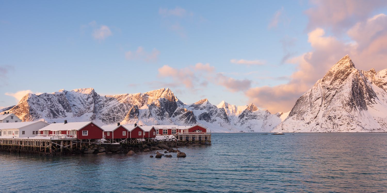 Classic red Norwegian rorbu fishing huts on stilts glow warmly beneath golden sunlit snow-capped Lofoten mountains.