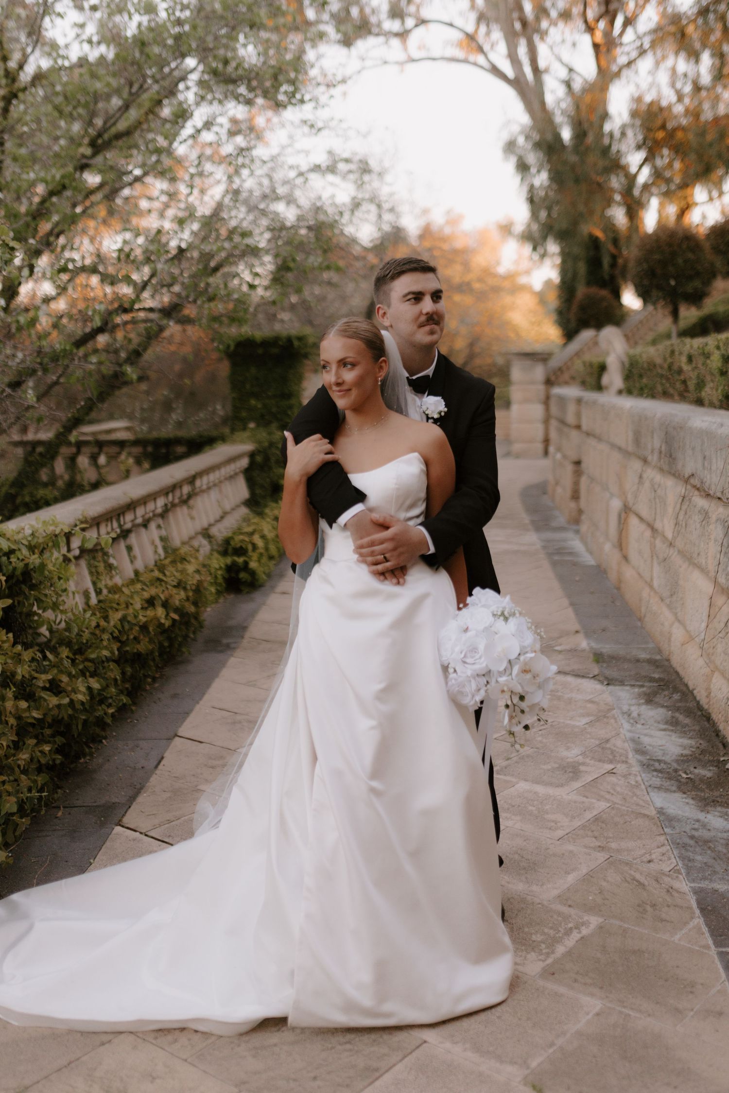 Bride and groom share a romantic kiss under a weeping willow tree by a lake, bride holding white bouquet.
