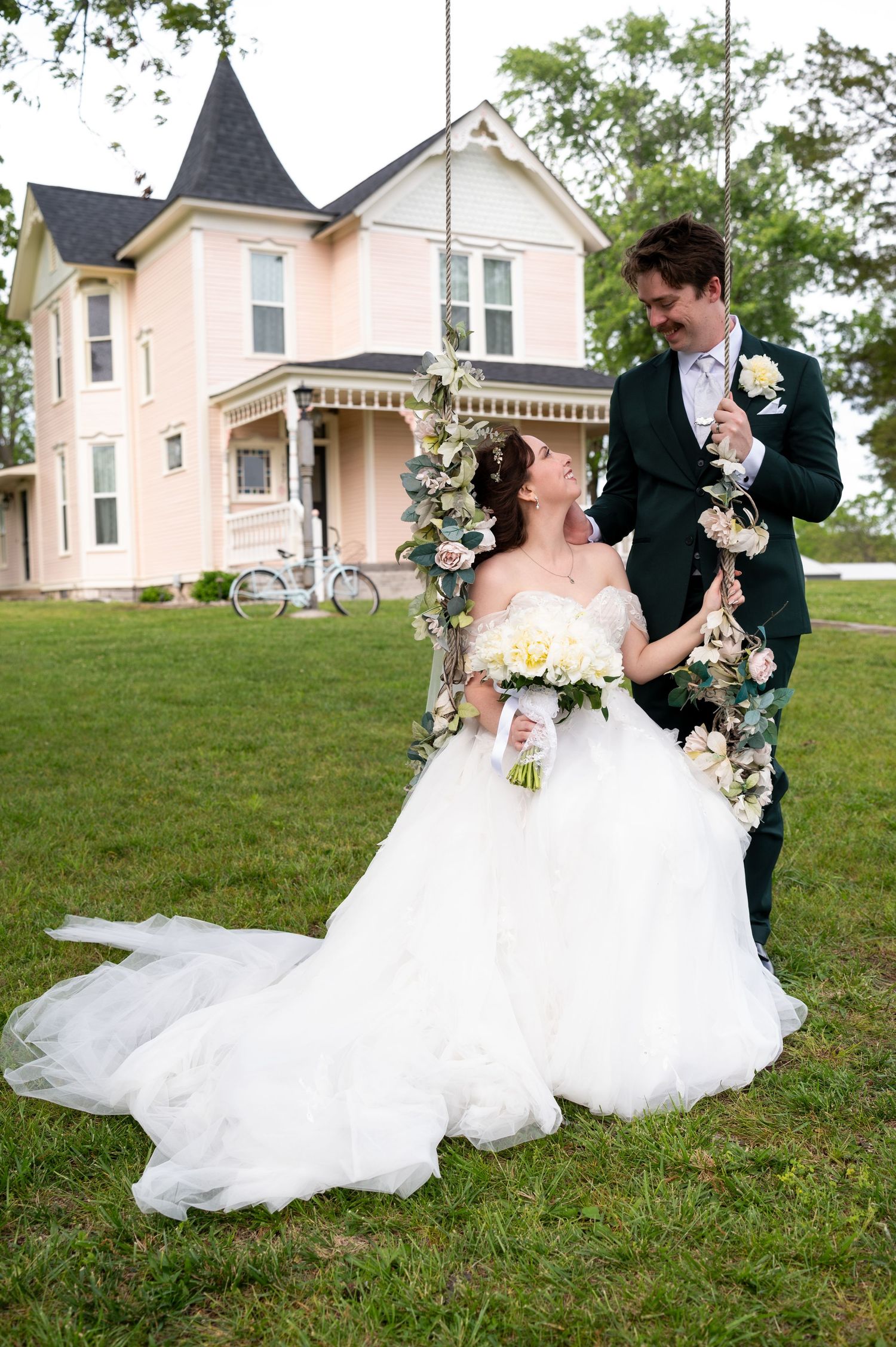 Bride in white gown sits on floral swing while groom in dark suit stands beside her, in front of a pink Victorian house.