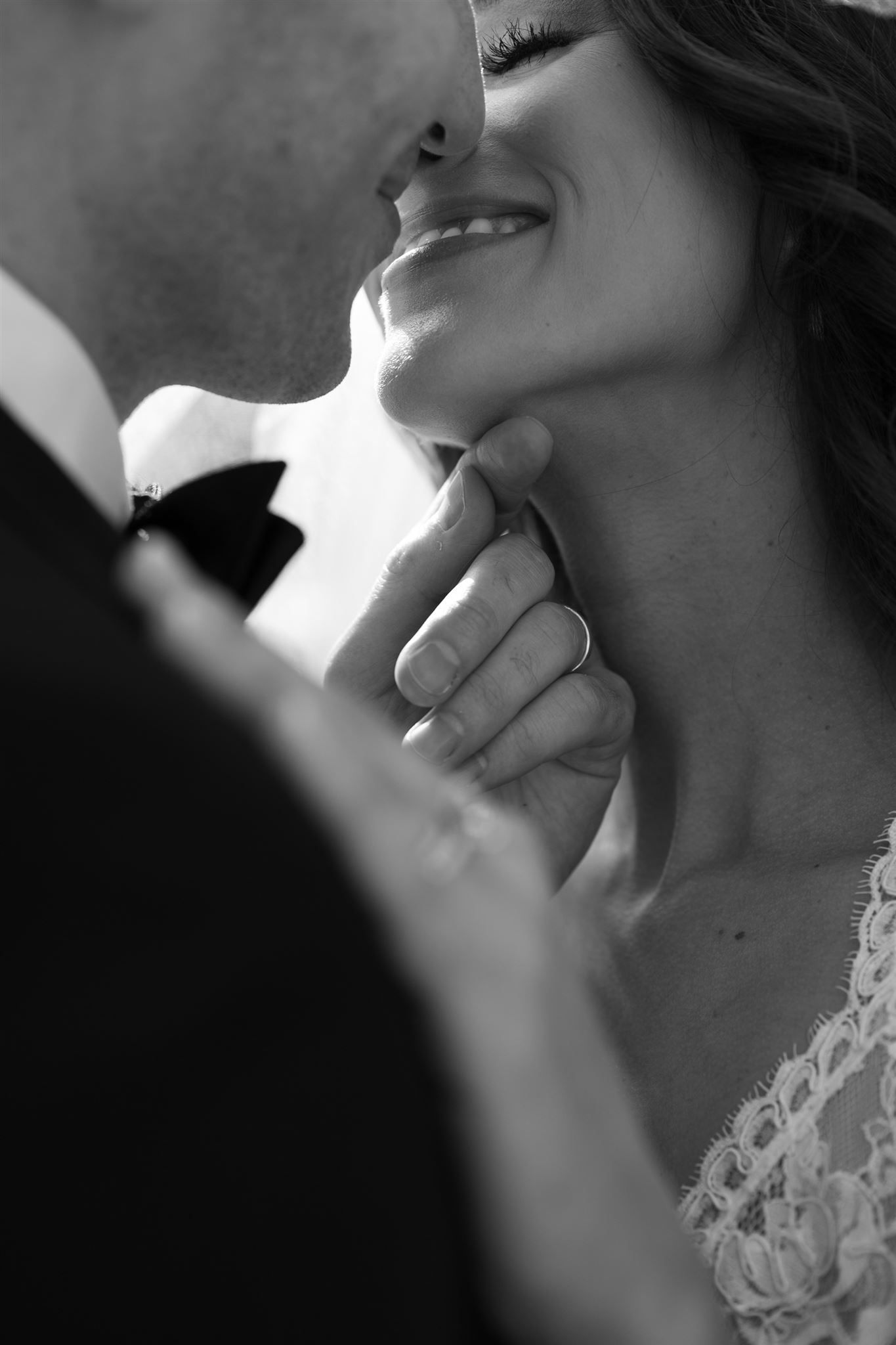 Black and white photo of a groom lifting bride's chin for a kiss, bride smiling in lace dress.