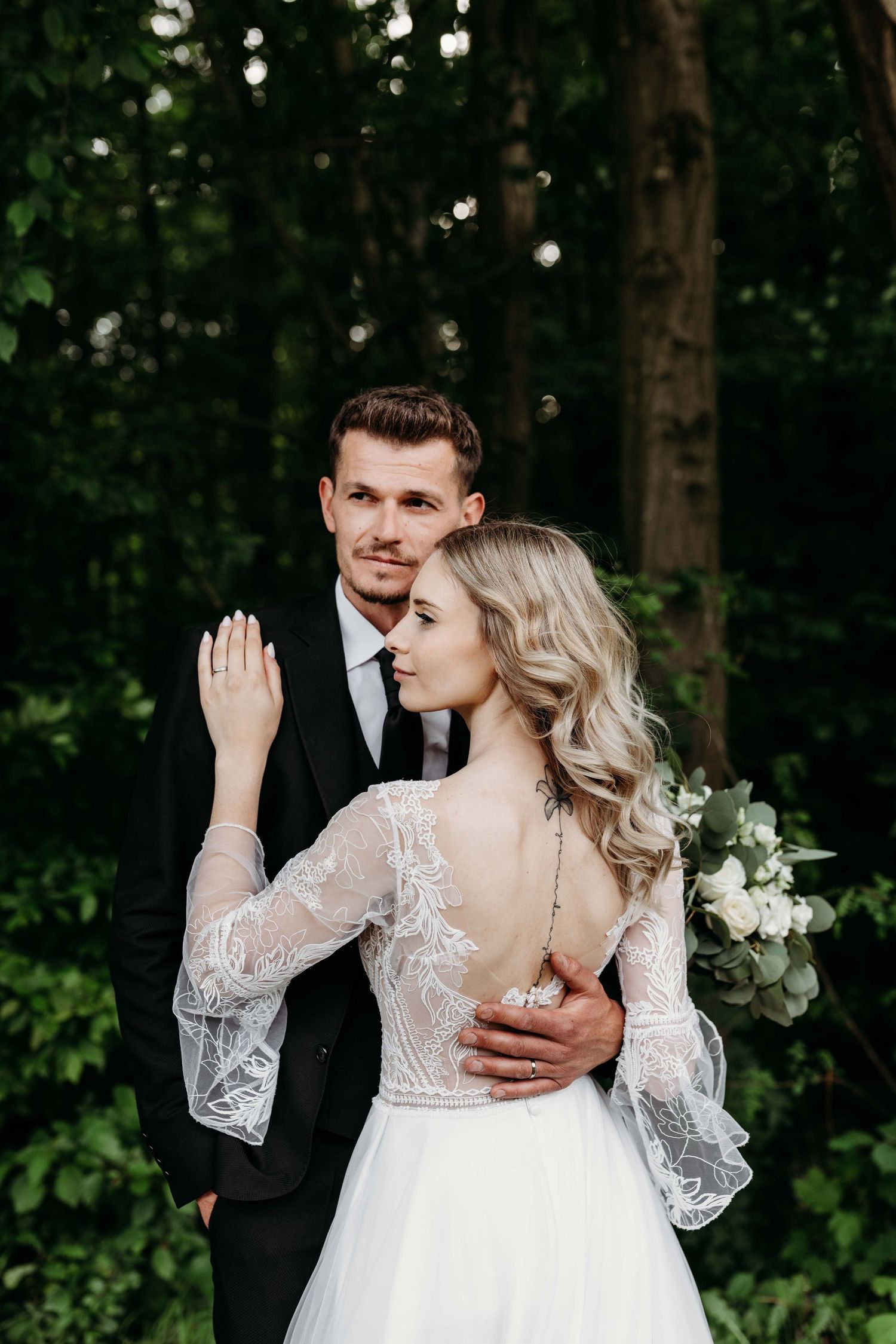 Bride in lace backless gown holds bouquet as groom in black suit embraces her in a lush green forest.