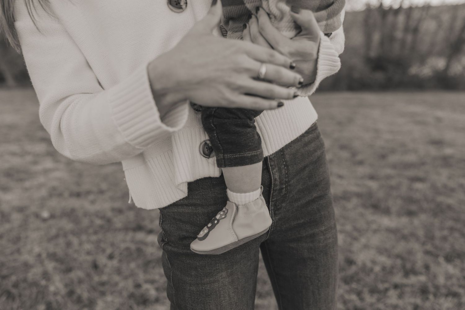 Black and white photo of a mother holding a baby in tiny booties outdoors on a grassy field.