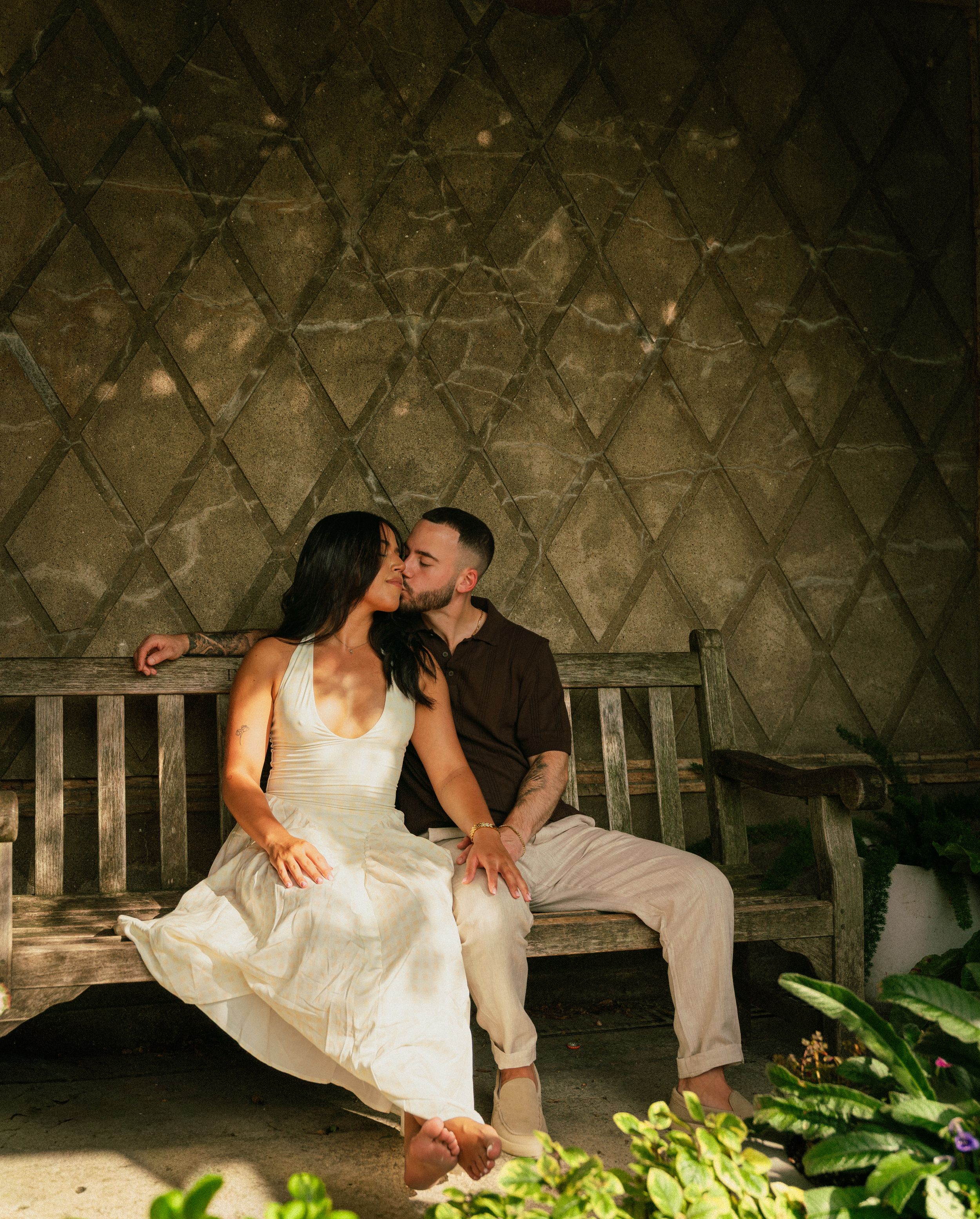 Couple sharing a romantic kiss on a wooden bench inside a yurt, woman in white dress, lush greenery surrounding them.