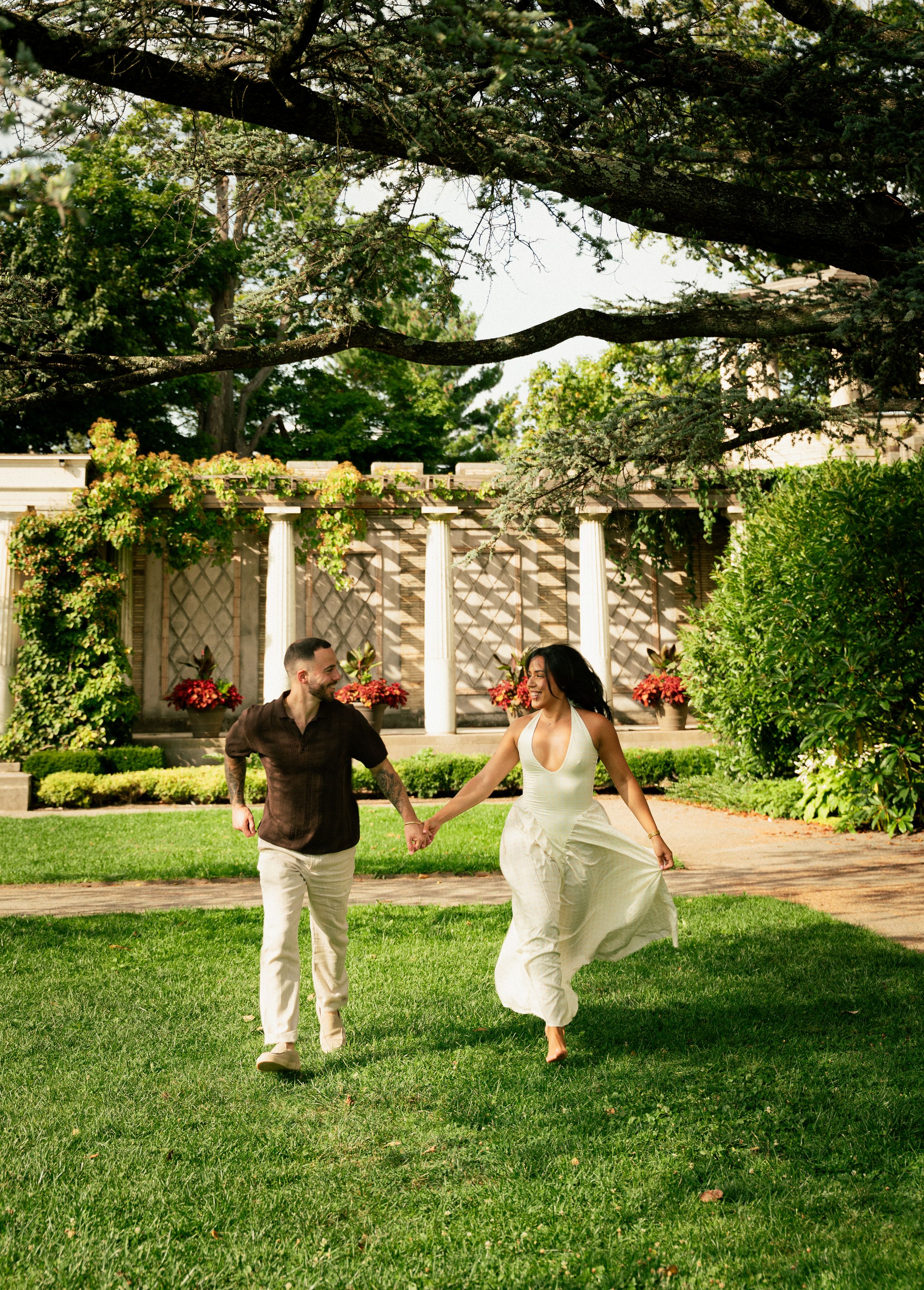 A couple holds hands and runs through a lush garden with columns and flowers in the background.