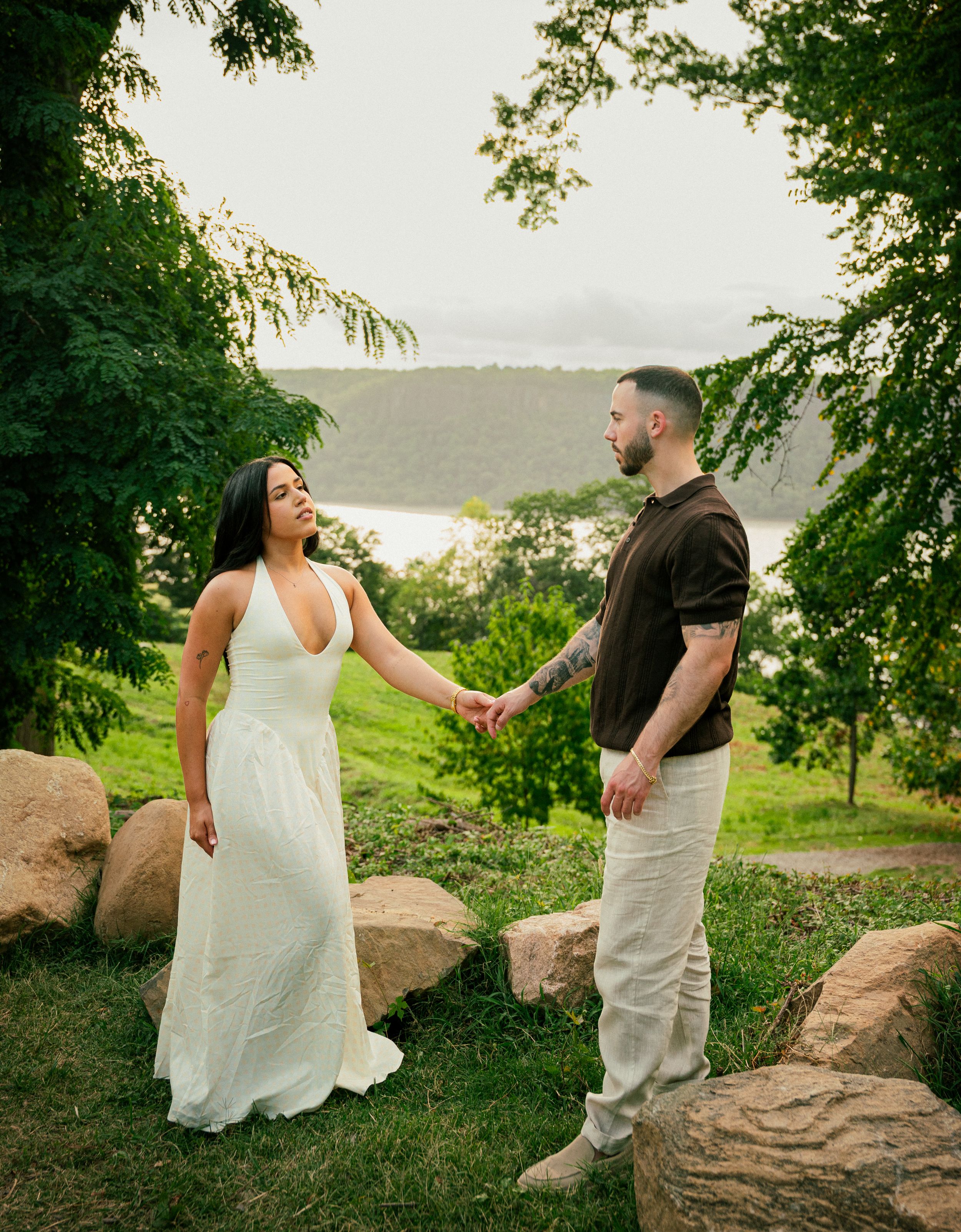 Couple holding hands in wedding attire outdoors, surrounded by lush greenery and rocks with a scenic water view.