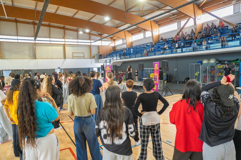 Large group of students gathered in a school gymnasium for a dance or sports event with spectators in the bleachers.