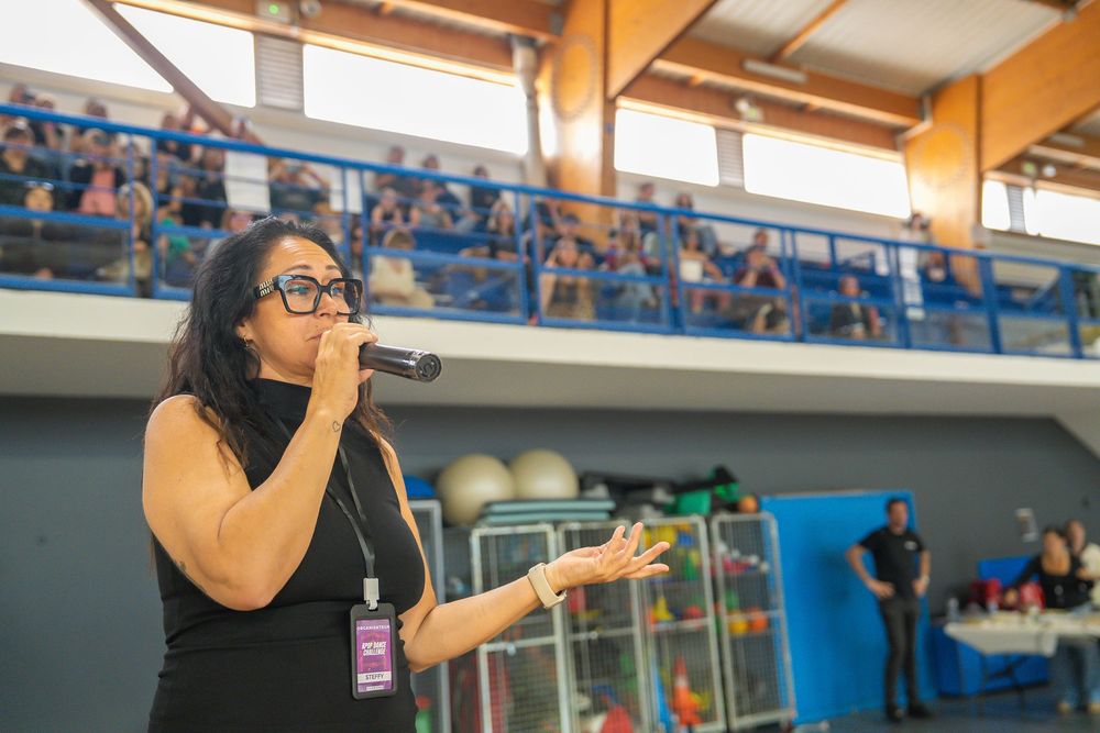 Woman speaking into microphone at indoor sports event with audience in bleachers behind her.