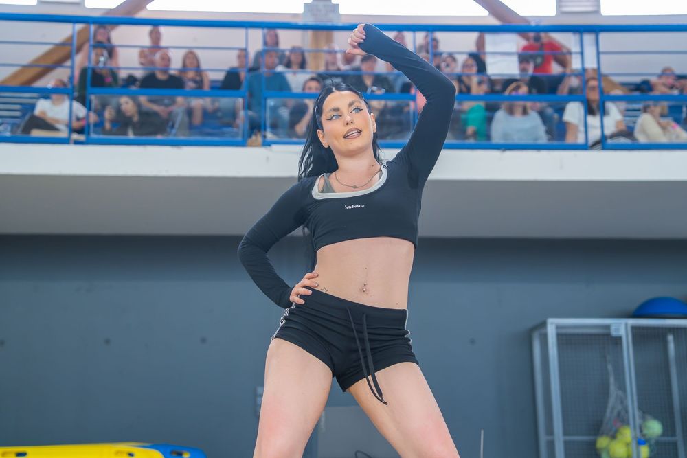A woman in a black crop top and shorts strikes a dynamic dance pose outdoors at a sports venue with spectators.