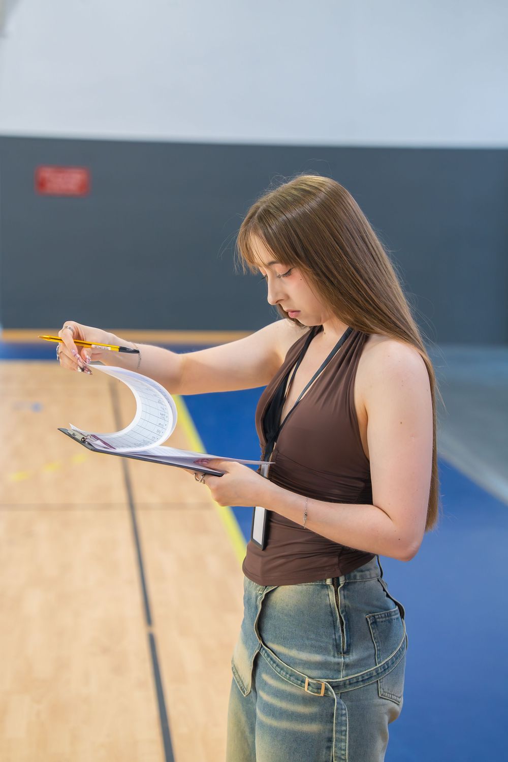 Young woman with long hair reviewing documents on a clipboard with a pencil in a gymnasium.