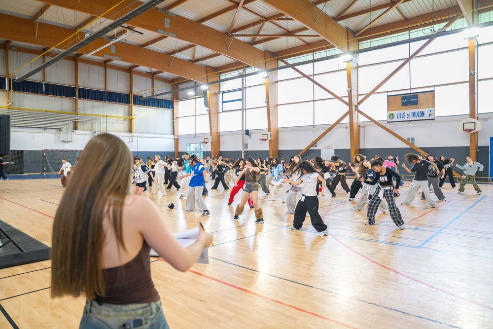 Large group dance class practicing choreography in a spacious gymnasium with wooden floors and high ceilings.