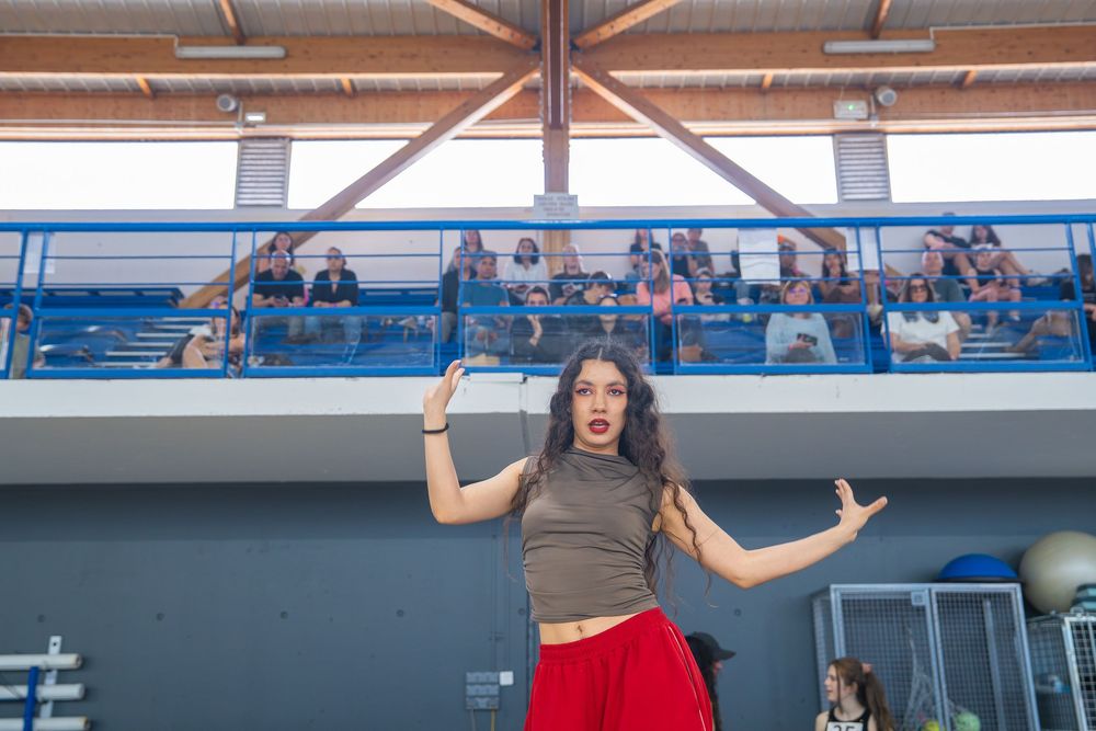 Young woman in red skirt and gray top dancing expressively indoors with audience watching from balcony.