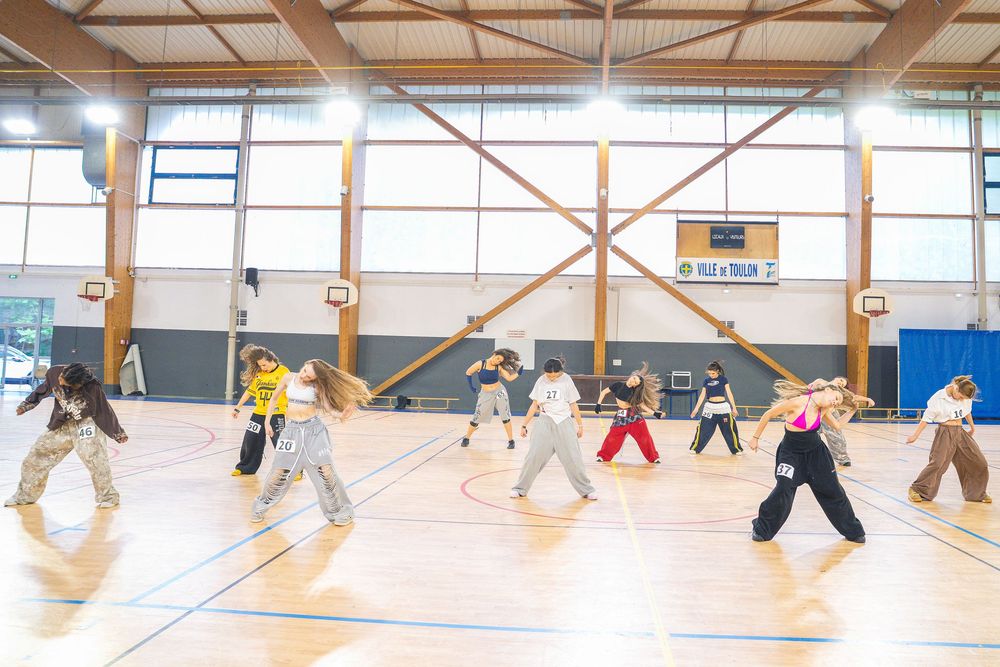 Group of dancers practicing hip-hop choreography in a gymnasium with wooden floors and high ceilings.