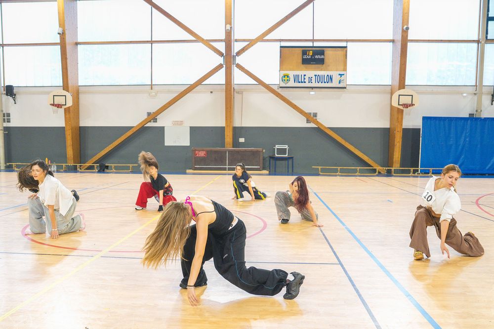 Young dancers practicing choreography on a gymnasium floor in an indoor sports hall with wooden flooring.