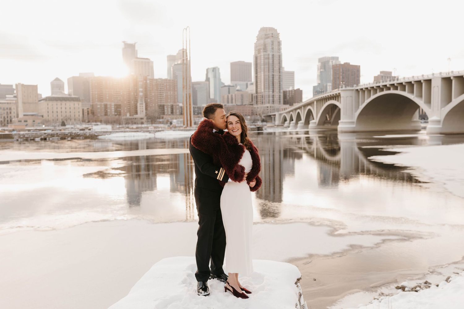 Couple embracing by a frozen river with a city skyline and bridge at sunset.