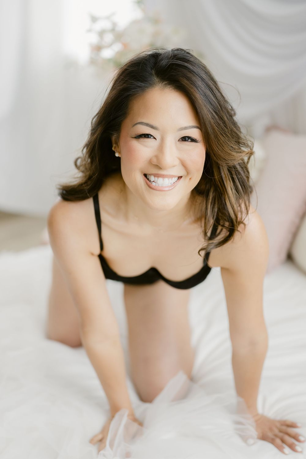 Smiling woman in black lingerie posing on a white bed in a bright, softly lit room.