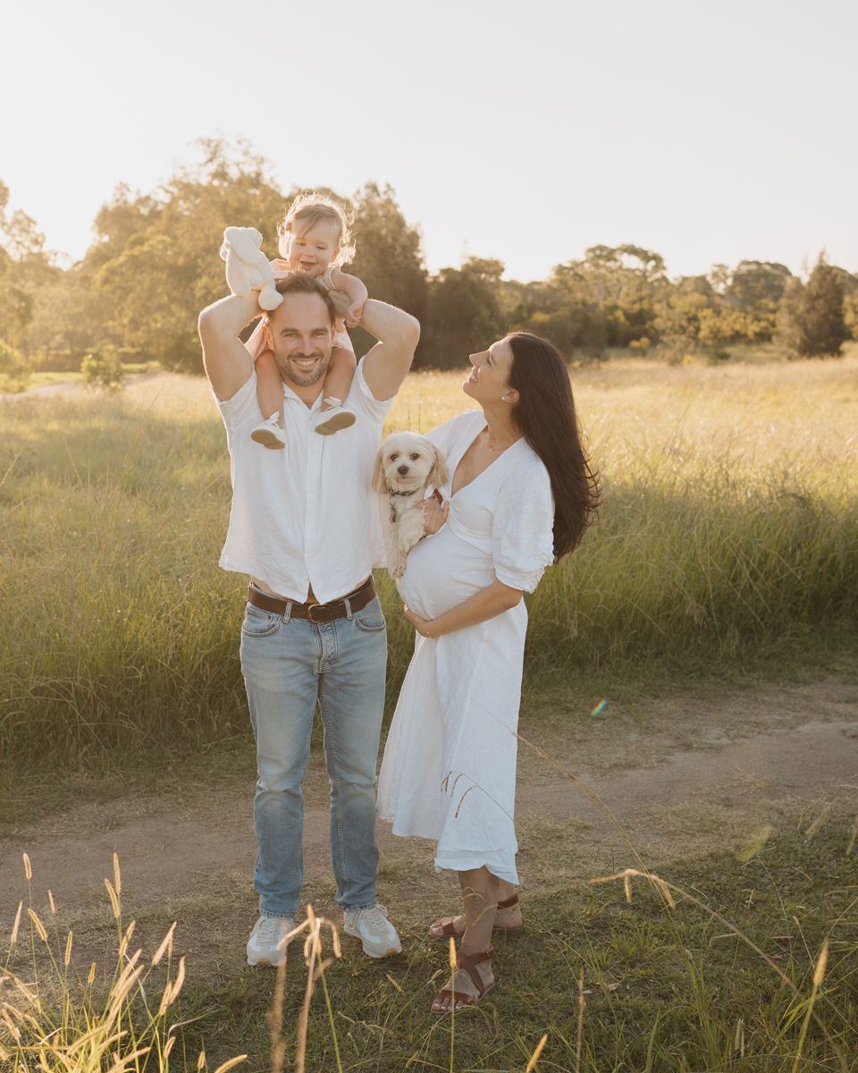 Happy family with toddler on dad's shoulders, holding a fluffy dog, posing in a sunny golden field in Rouse hill regional park photoshoot