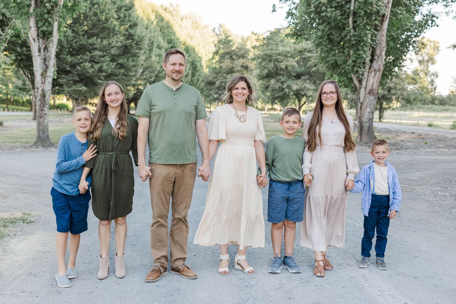 Family of seven smiling outdoors on a sunny day, posing together on a gravel path surrounded by green trees.