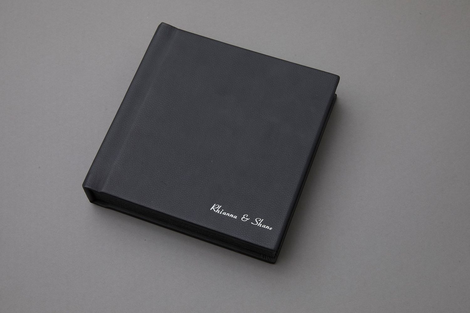 Black leather wedding photo album with silver cursive names embossed on cover, displayed on gray background.