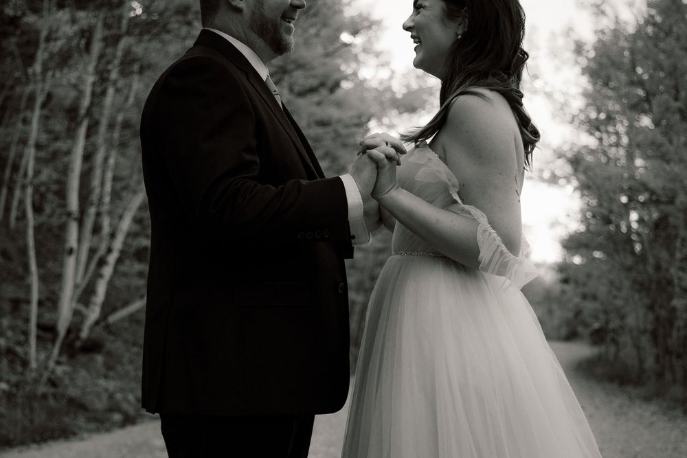 Black and white photo of a bride and groom holding hands and laughing together on a tree-lined path.