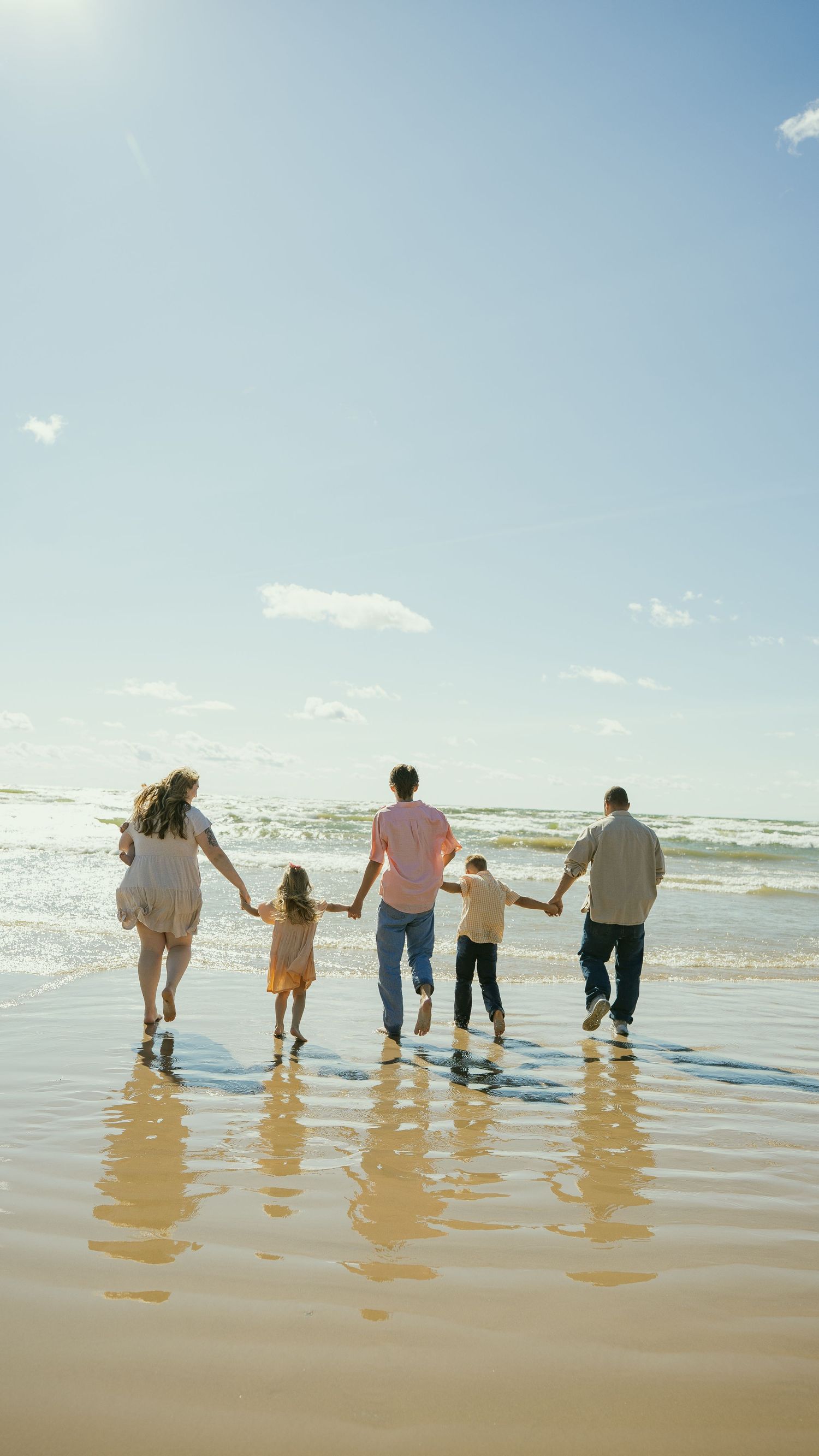 Family of five holding hands walking toward ocean waves on a sunny sandy beach.