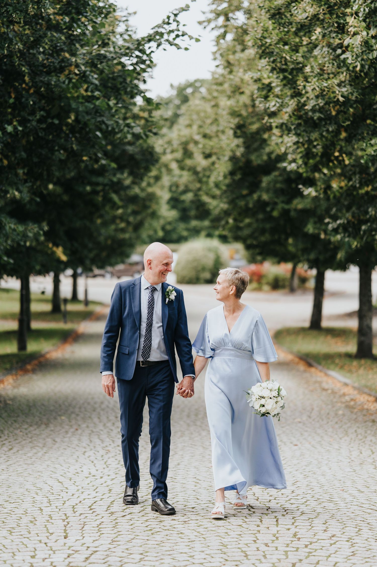 Elegant mature couple standing on a wooden garden bridge surrounded by lush greenery, dressed in wedding attire.