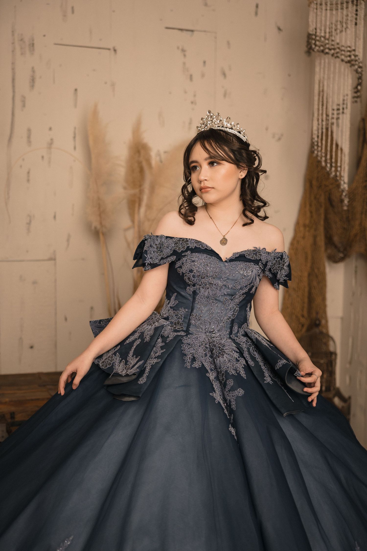 Young woman in navy blue ball gown with lace details and silver tiara poses elegantly in a rustic studio setting.