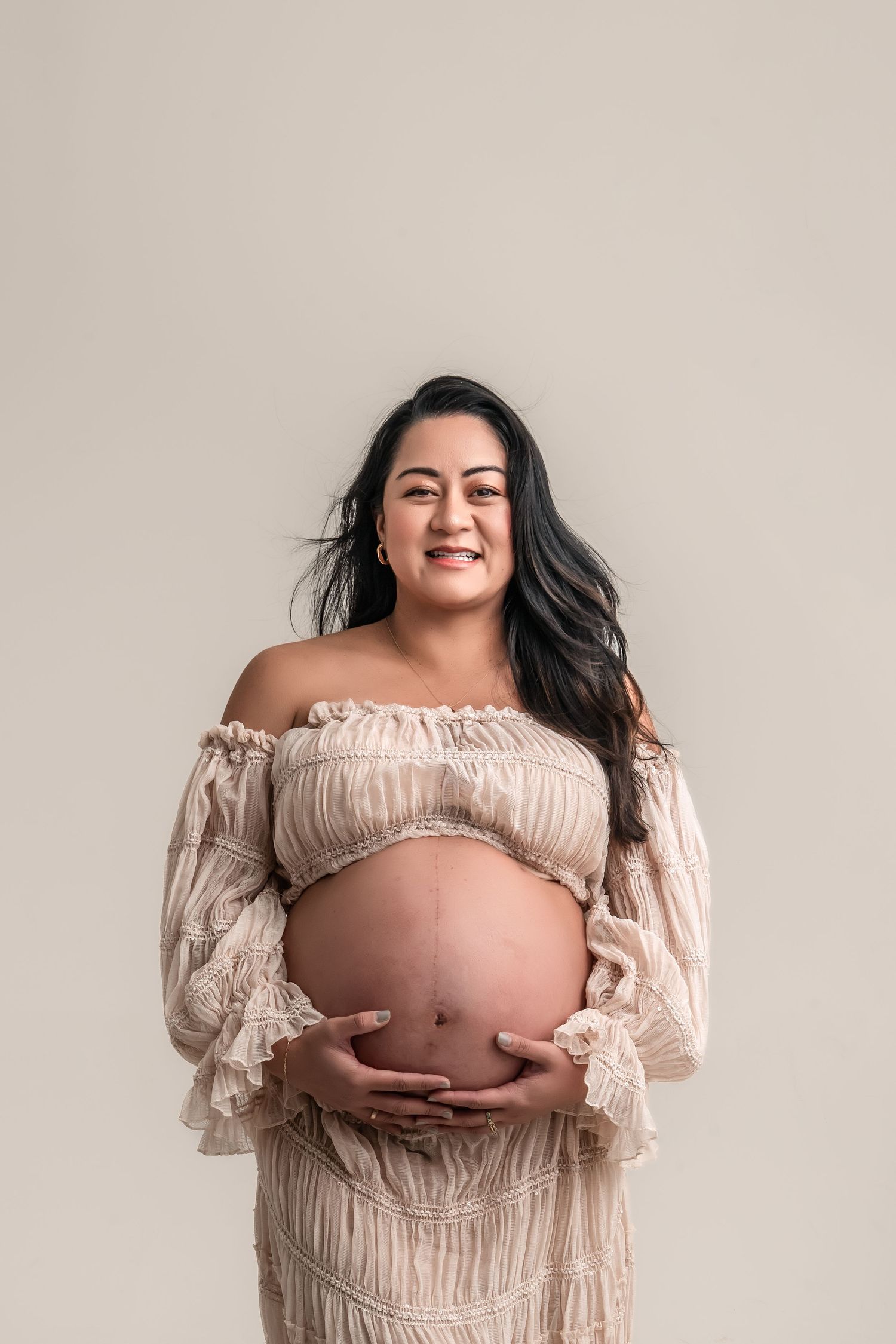 Smiling pregnant woman in off-shoulder cream ruffled dress cradling baby bump against neutral background.
