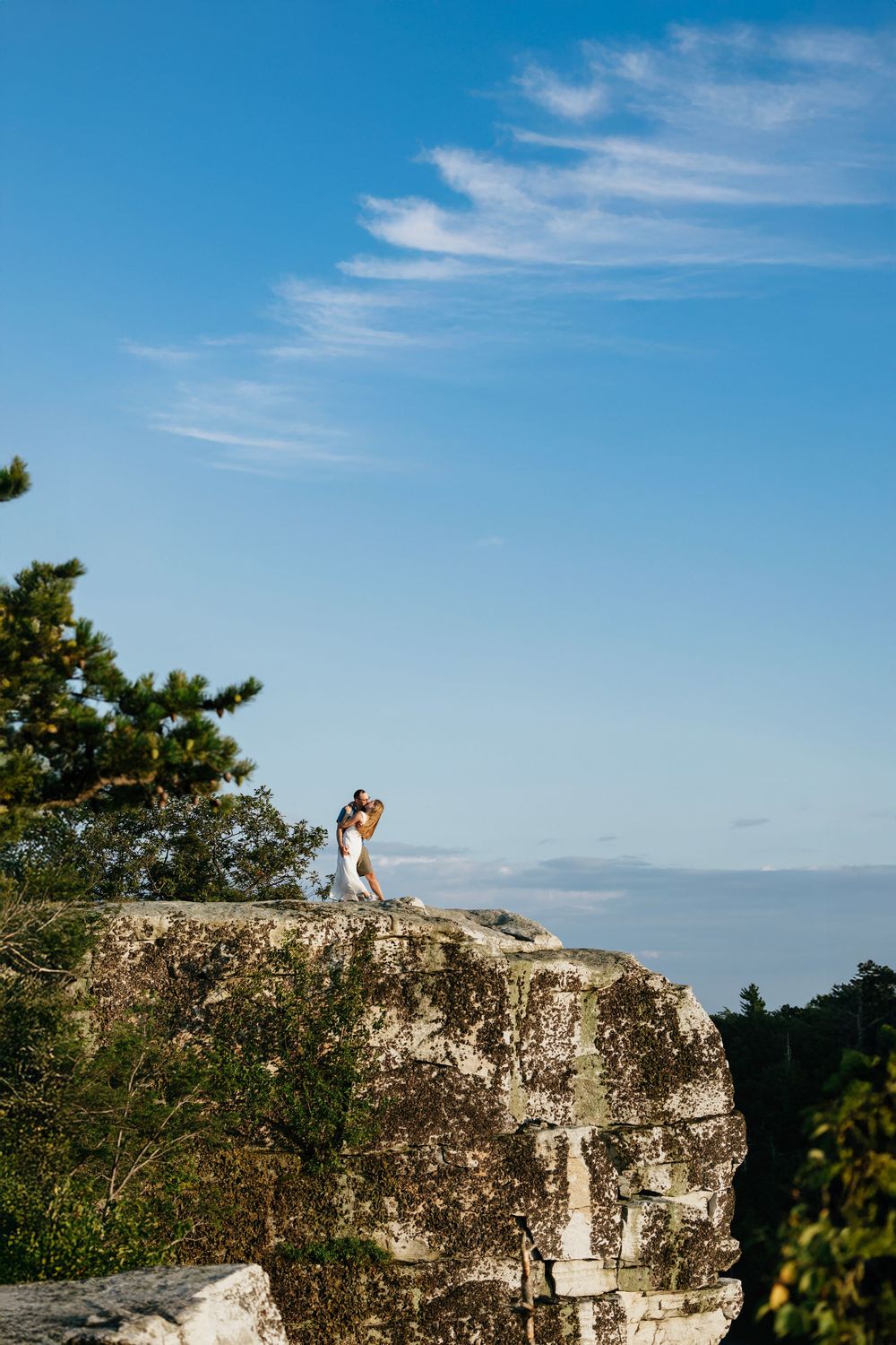 Person in white outfit standing on rocky cliff edge against blue sky with wispy clouds and green trees.