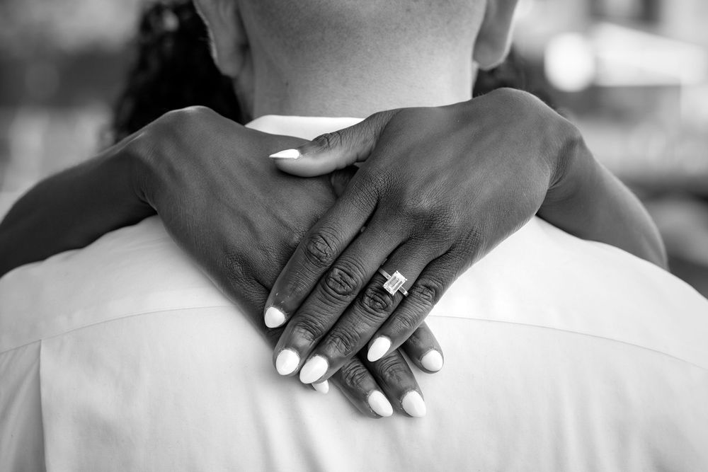 Black and white photo of a couple embracing, showing hands with a diamond engagement ring and white manicured nails.