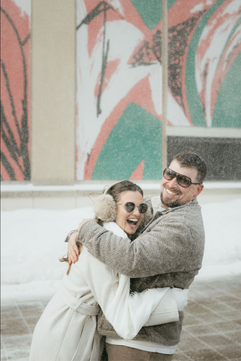 Couple hugging and laughing in snow wearing sunglasses, white coat, and gray jacket in front of colorful mural.