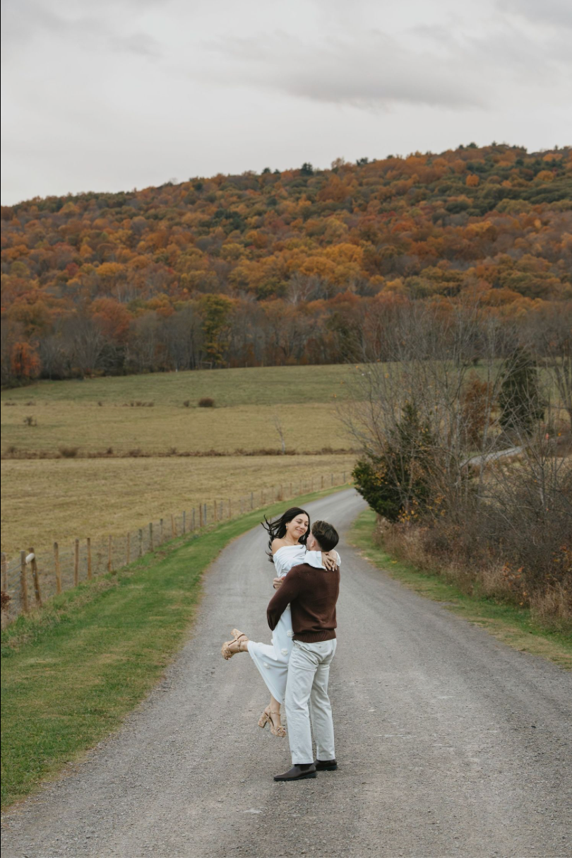 Couple embracing on a rural road surrounded by vibrant autumn foliage and green fields under an overcast sky.