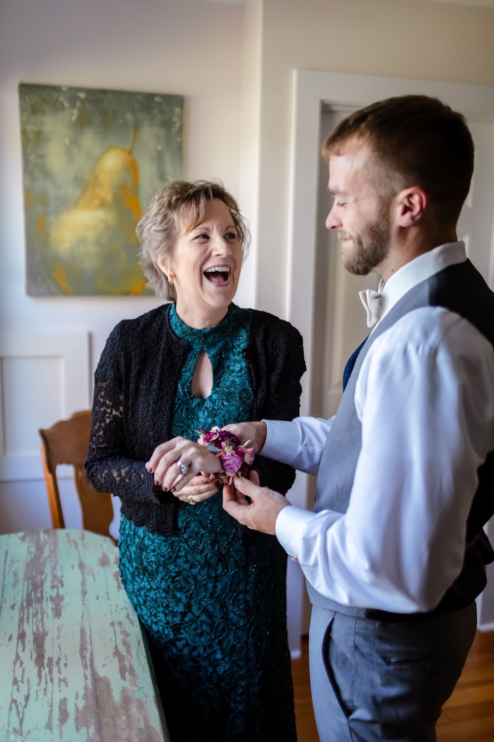 Mother of groom laughs while he puts on her corsage in a white walled room with green and yellow painting of pear on wall at Riverledge Farm