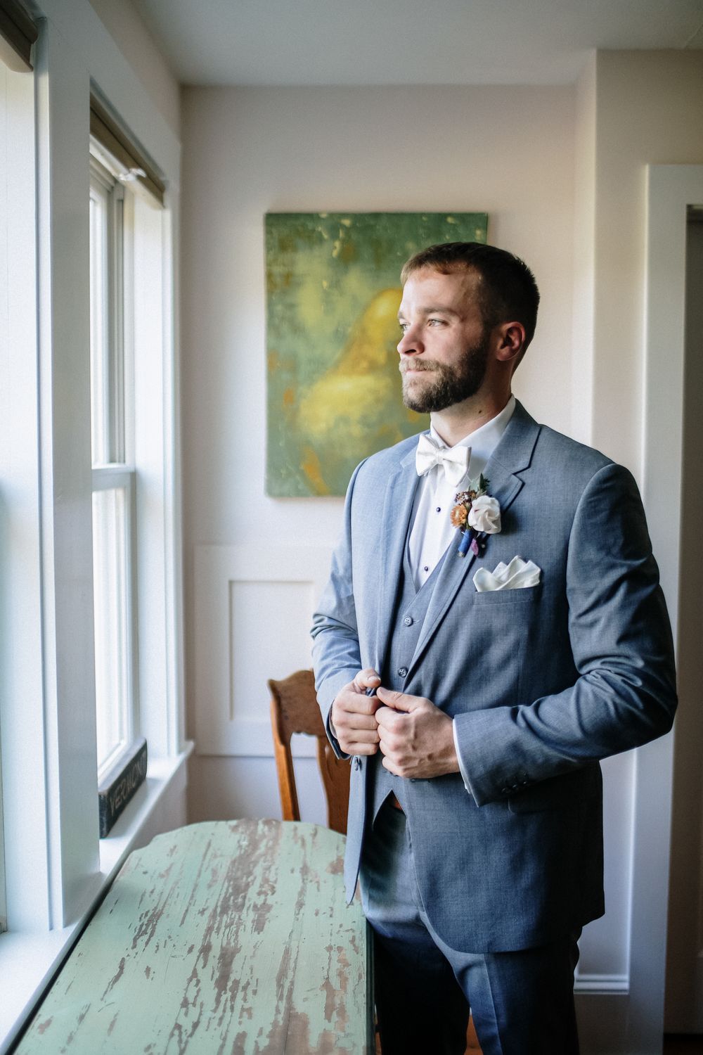 Groom in gray suit with boutonniere holds jacket to self near window with natural light for getting ready photos at Grafton, VT wedding.